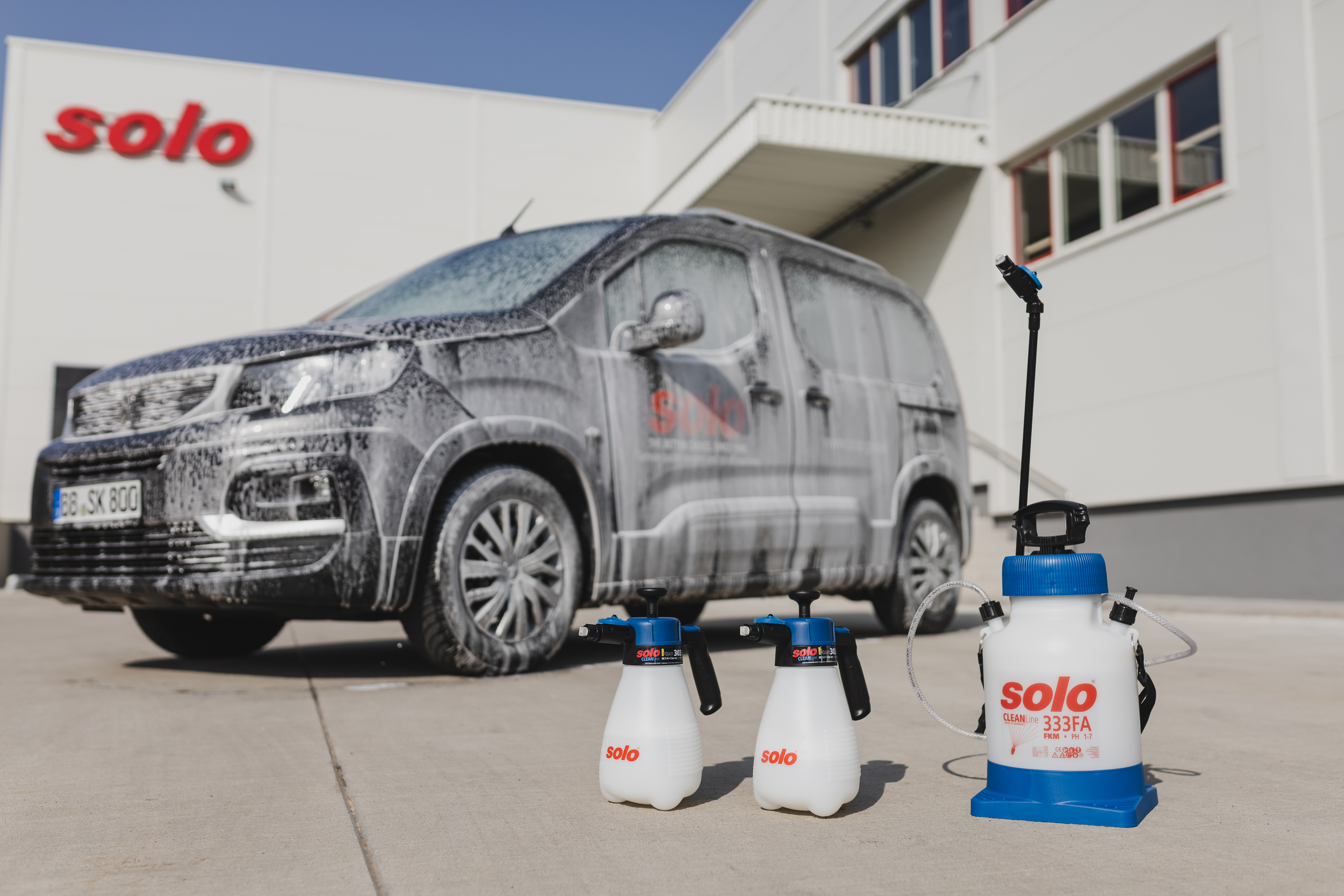 Mit Schaum bedecktes Firmenfahrzeug vor dem SOLO-Gebäude, im Vordergrund stehen die Sprühgeräte SOLO 301 FA, 302 FA und 333 FA für professionelle Fahrzeugreinigung.